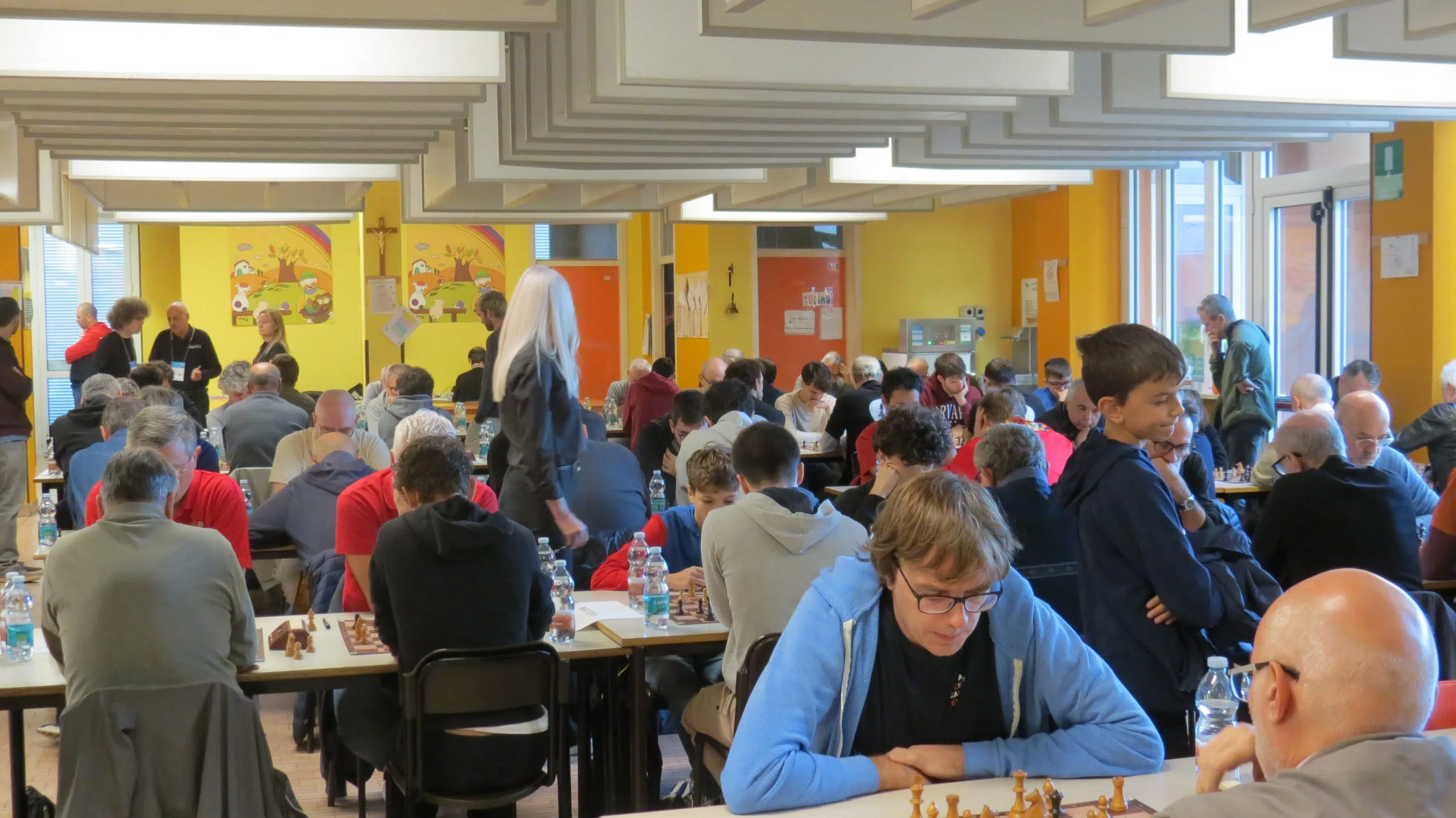 A crowded chess event with many participants seated at tables, engaged in games. The room has bright colors and artwork on the walls.