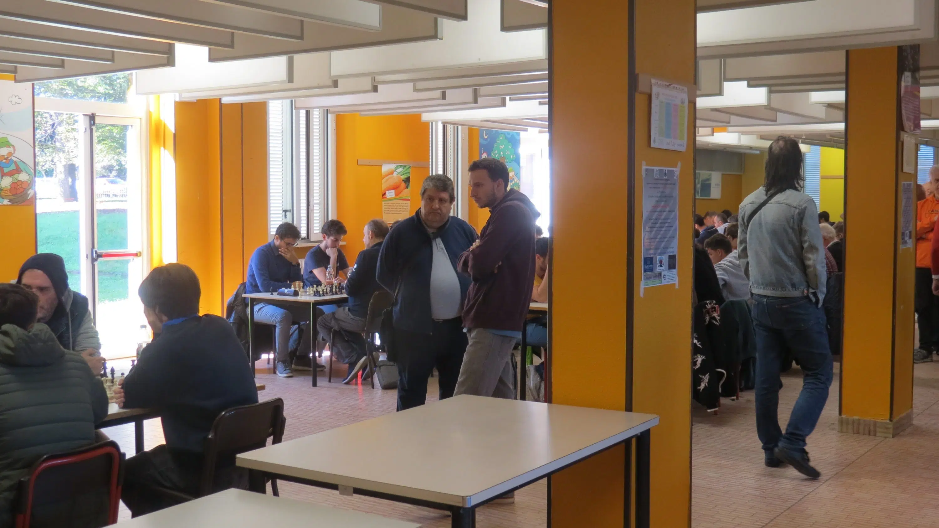 A busy cafeteria with students sitting at tables, some talking and others walking around.
