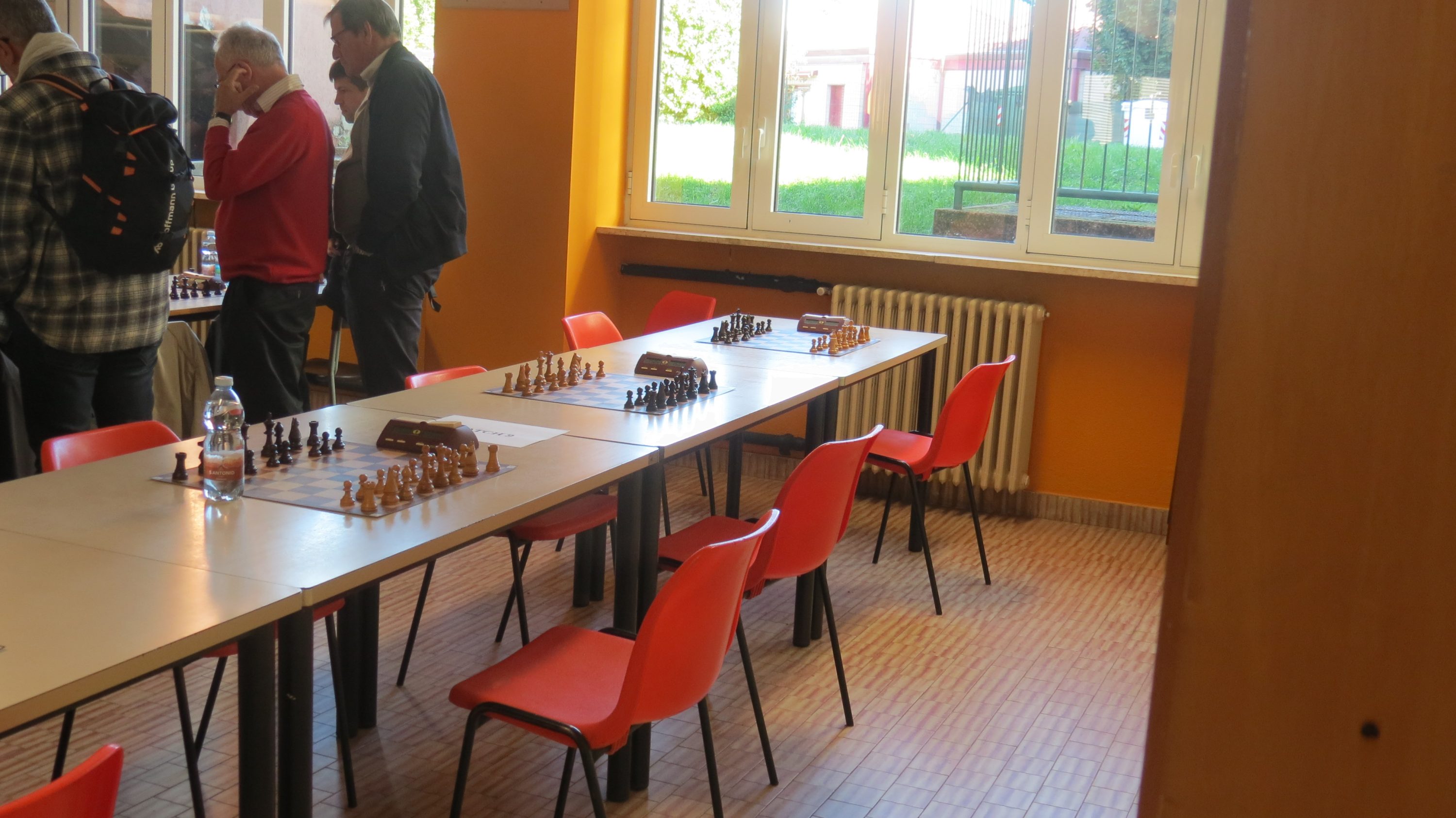 A room featuring a long table set up for a chess game, accompanied by several orange chairs and large windows providing natural light.