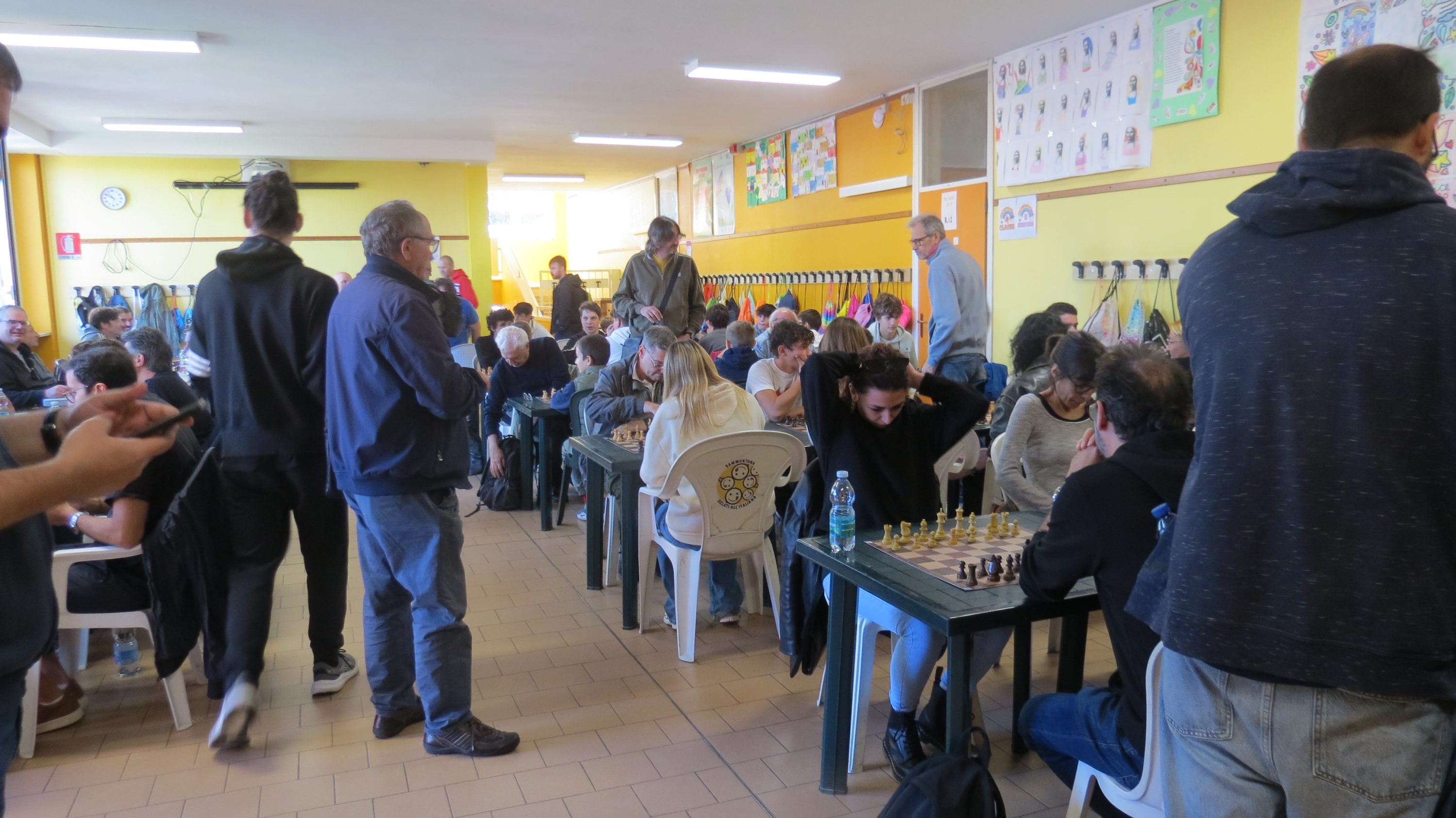 A crowded room filled with people playing chess at tables, with yellow walls and posters in the background.