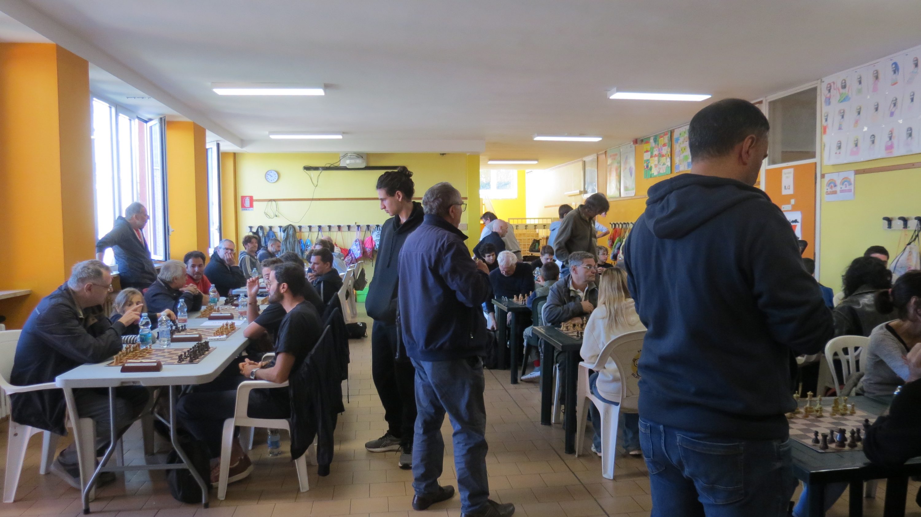 Una sala llena de personas jugando al ajedrez en mesas, con paredes amarillas y una atmósfera animada.
