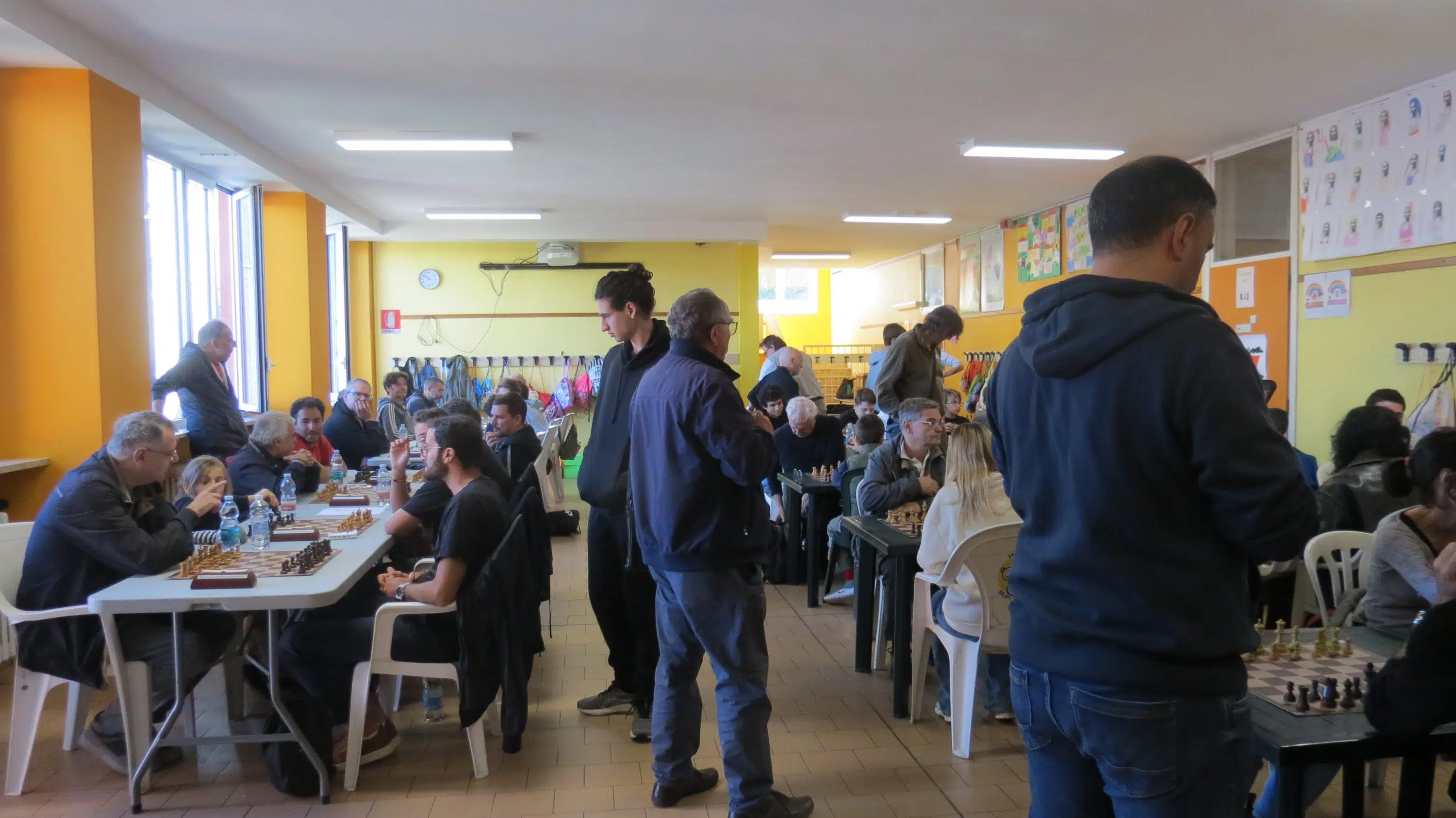 Una sala llena de personas jugando al ajedrez en mesas, con paredes amarillas y una atmósfera animada.