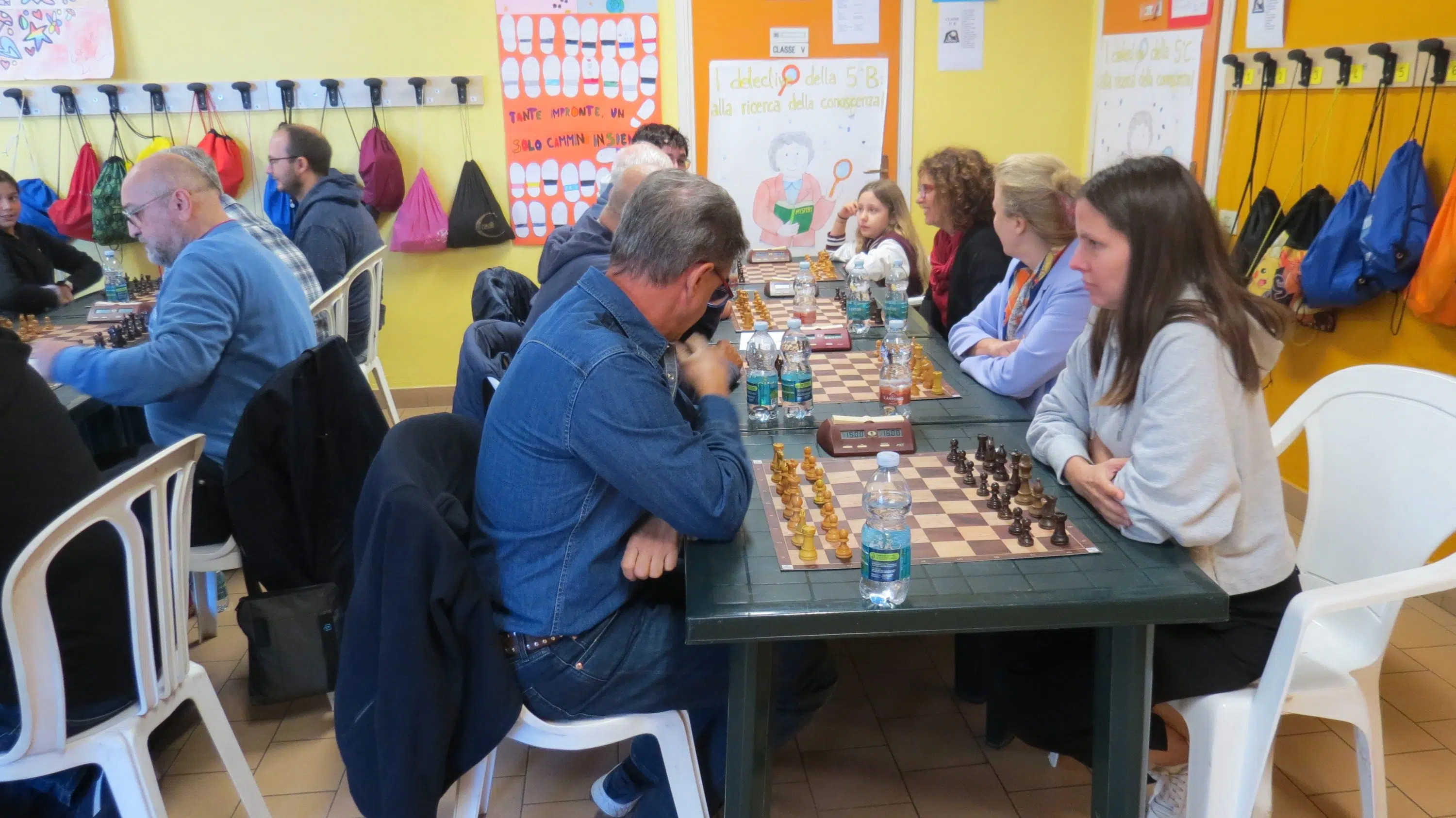 Mature individuals engaged in a chess game at a table, with a colorful backdrop of a room filled with backpacks and posters.