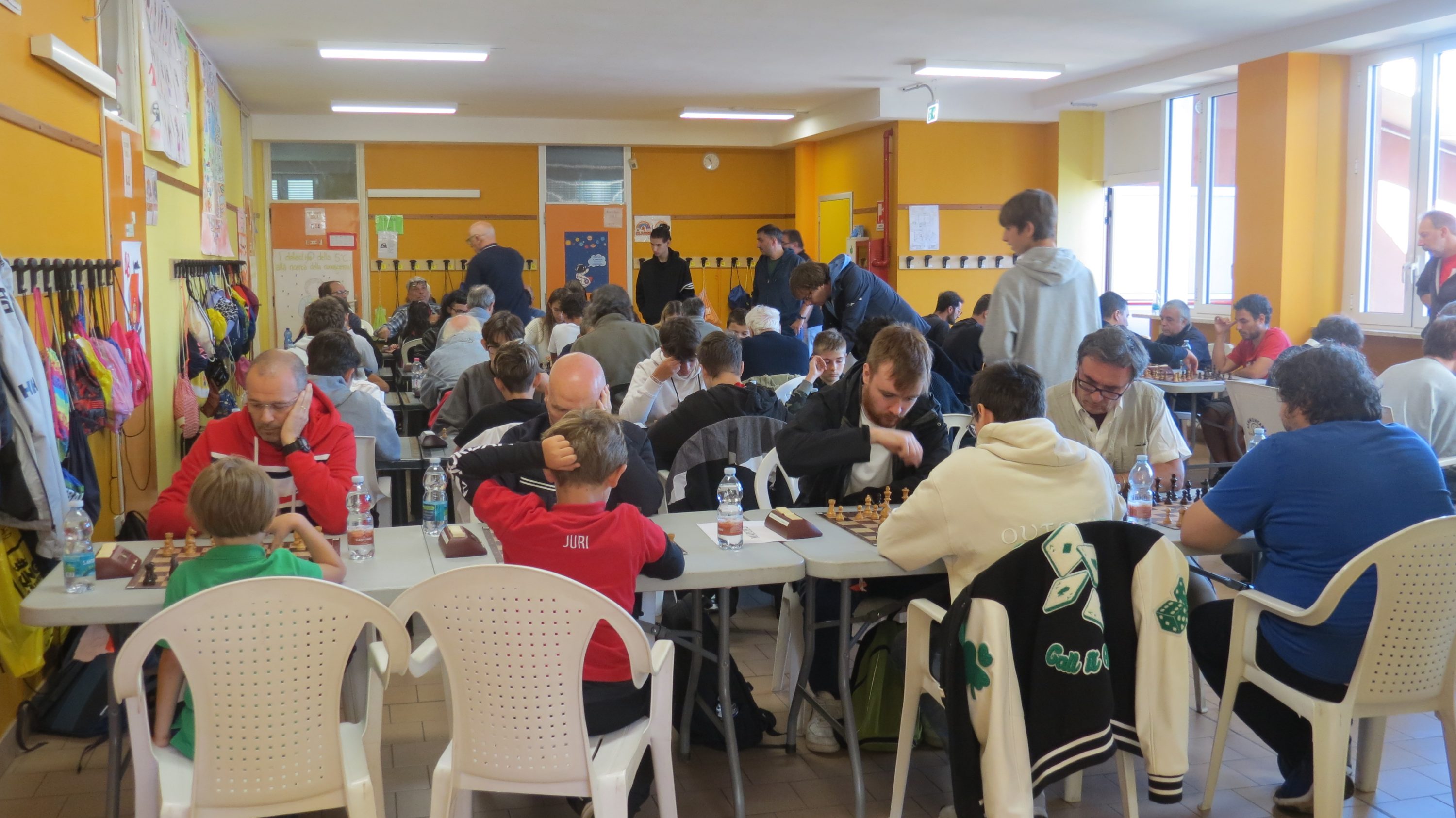 Una sala llena de gente sentada en mesas, participando en una actividad grupal. Las paredes son de color naranja y hay varios niños y adultos en la escena.