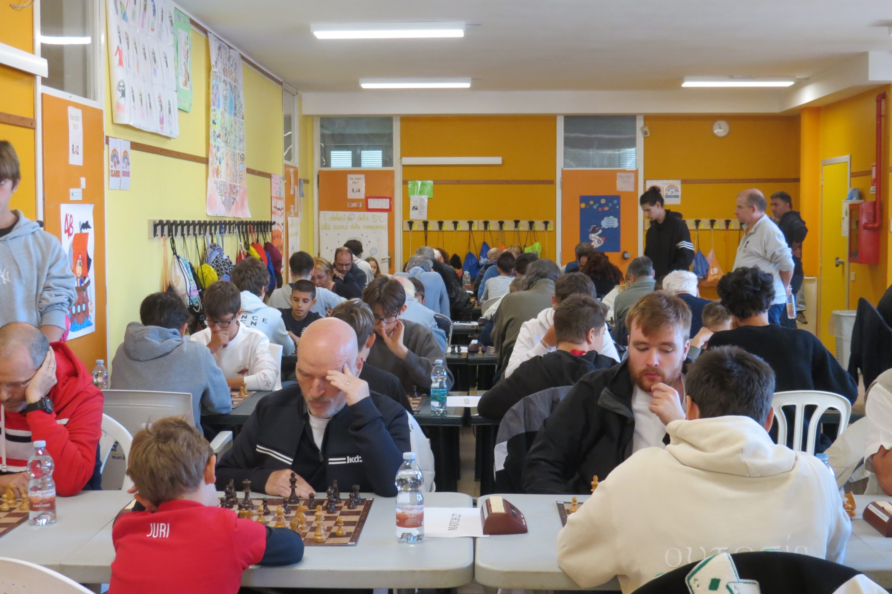 Una sala de juegos con participantes de un torneo de ajedrez sentados en mesas, concentrados en sus partidas. Las paredes son de color amarillo y hay carteles educativos en el fondo.
