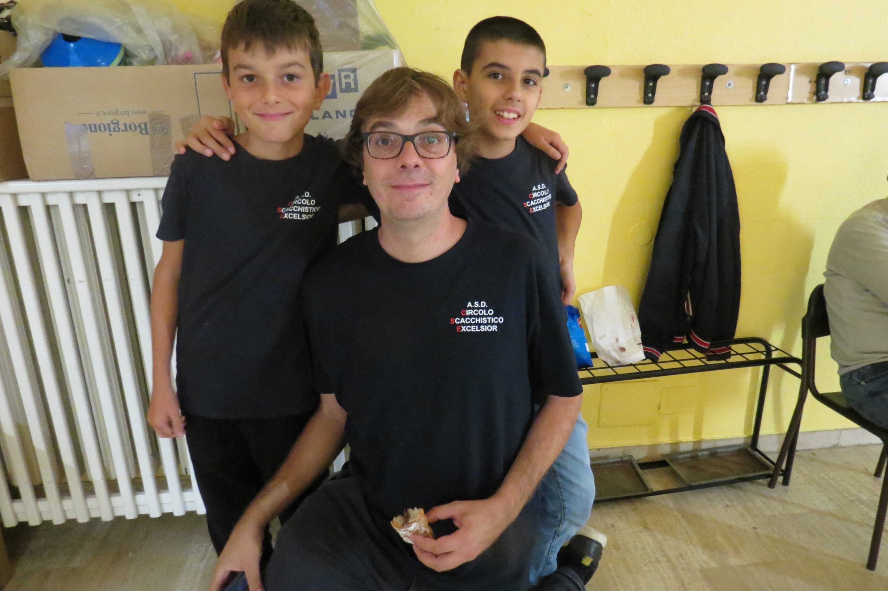 Un adulto sentado con dos niños, todos sonrientes, en un entorno interior con paredes amarillas y objetos en segundo plano.