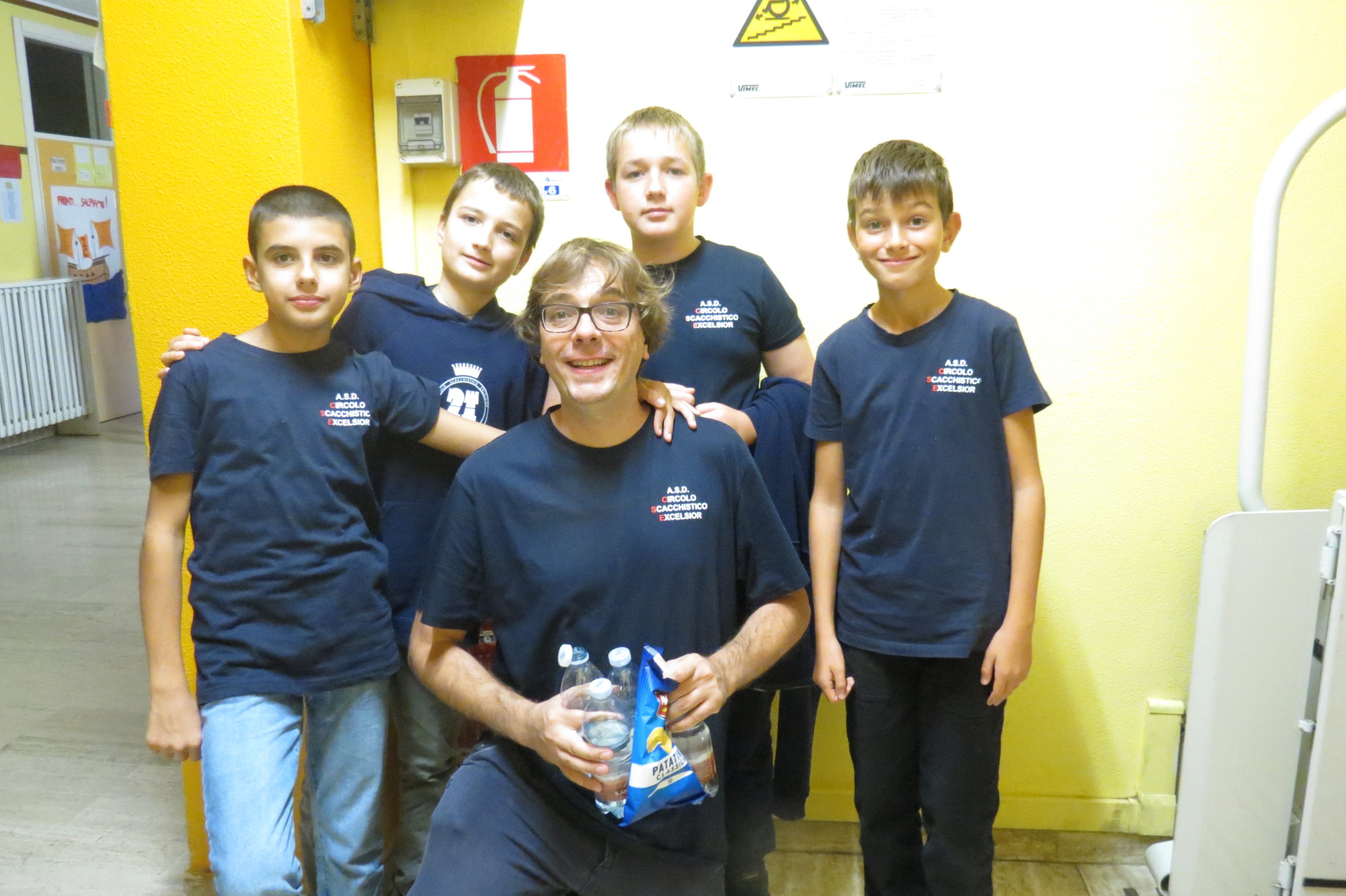 Un grupo de cinco jóvenes posando juntos con camisetas oscuras, sonriendo y sosteniendo botellas de agua en un entorno interior con paredes amarillas.