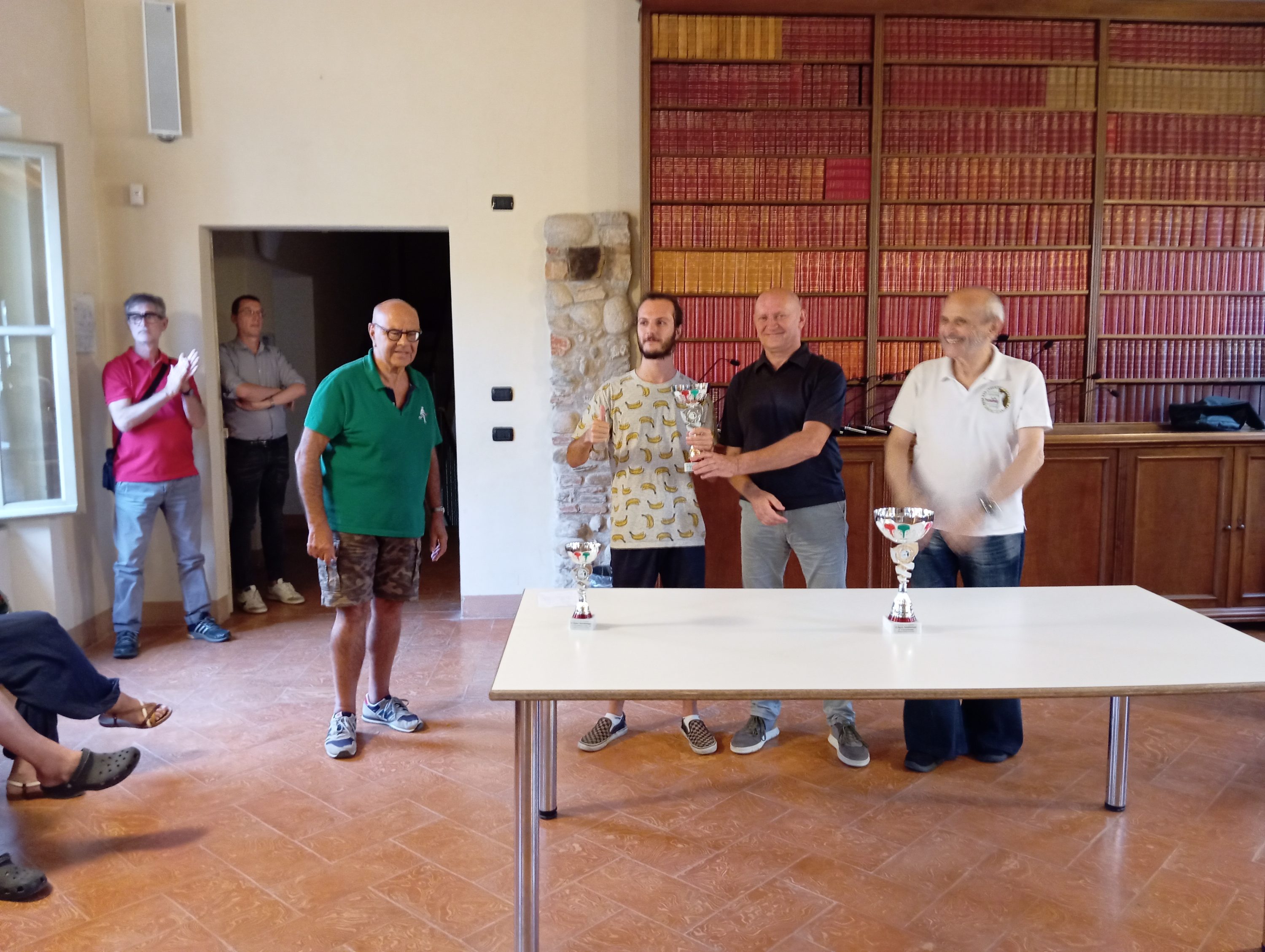 Cerimonia di premiazione del Torneo Semilampo con quattro partecipanti, ognuno dei quali riceve una coppa, in una sala consiliare arredata con libri.