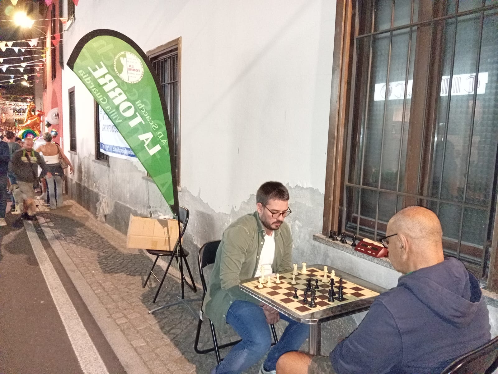 Due uomini giocano a scacchi all'aperto mentre passanti osservano l'attività.