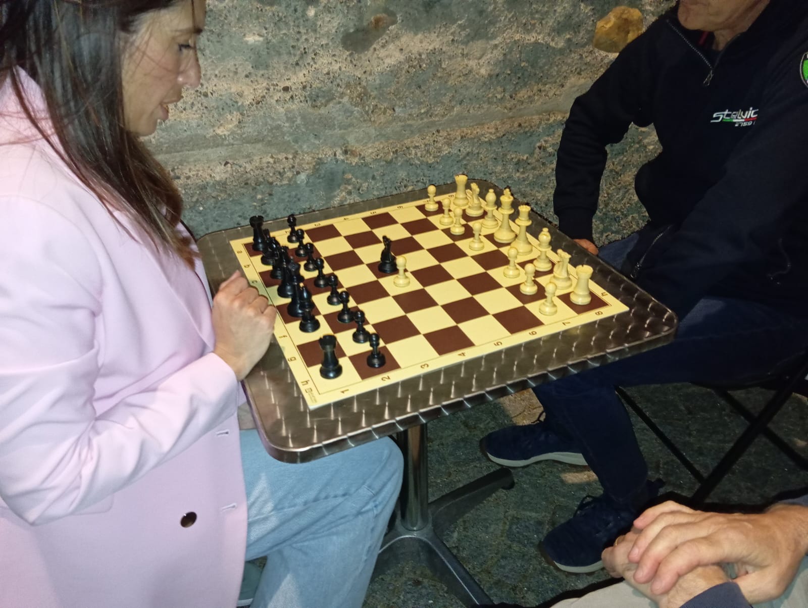 Una donna che gioca a scacchi su un tavolo con una scacchiera in legno, mentre altre persone sono sedute vicino a lei.