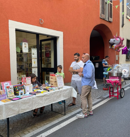 Un momento dell'iniziativa organizzata dalla Pro Loco di Binago, con un tavolo espositivo e visitatori che interagiscono, in un contesto urbano caratterizzato da edifici colorati.