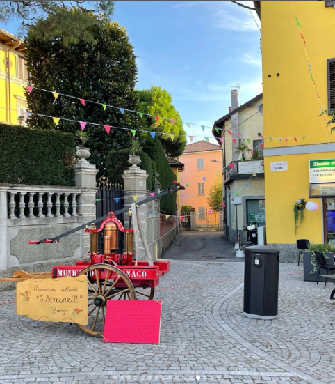 Piazza di Binago con un carretto rosso decorato e legato, circondato da alberi e bandierine colorate.
