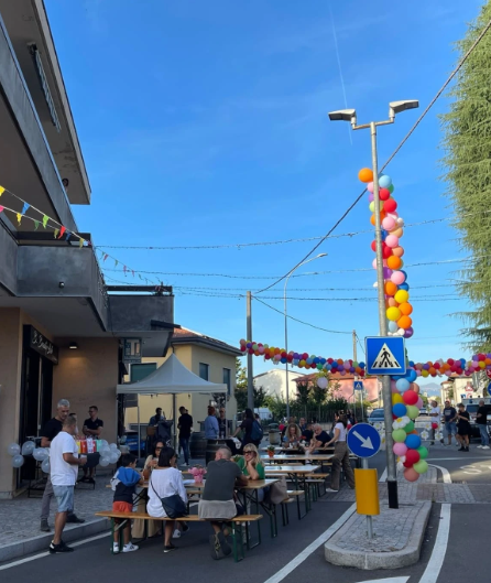 Evento all'aperto con tavoli disposti lungo la strada, decorati con palloncini colorati. Persone sedute e in movimento, con un'atmosfera festosa sotto un cielo sereno.