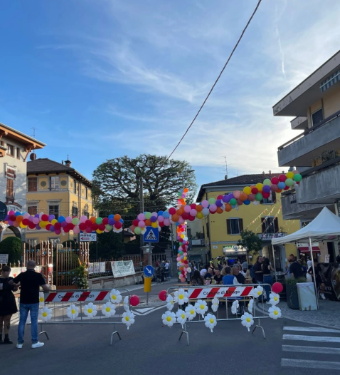 Vista di un evento all'aperto con decorazioni colorate, persone che partecipano sotto un arco di palloncini, e stendardi in una strada di Binago.