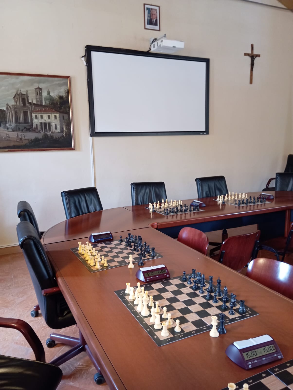 Un tavolo di legno con scacchiere pronte per il torneo, in una sala con sedie nere e un proiettore sullo sfondo.