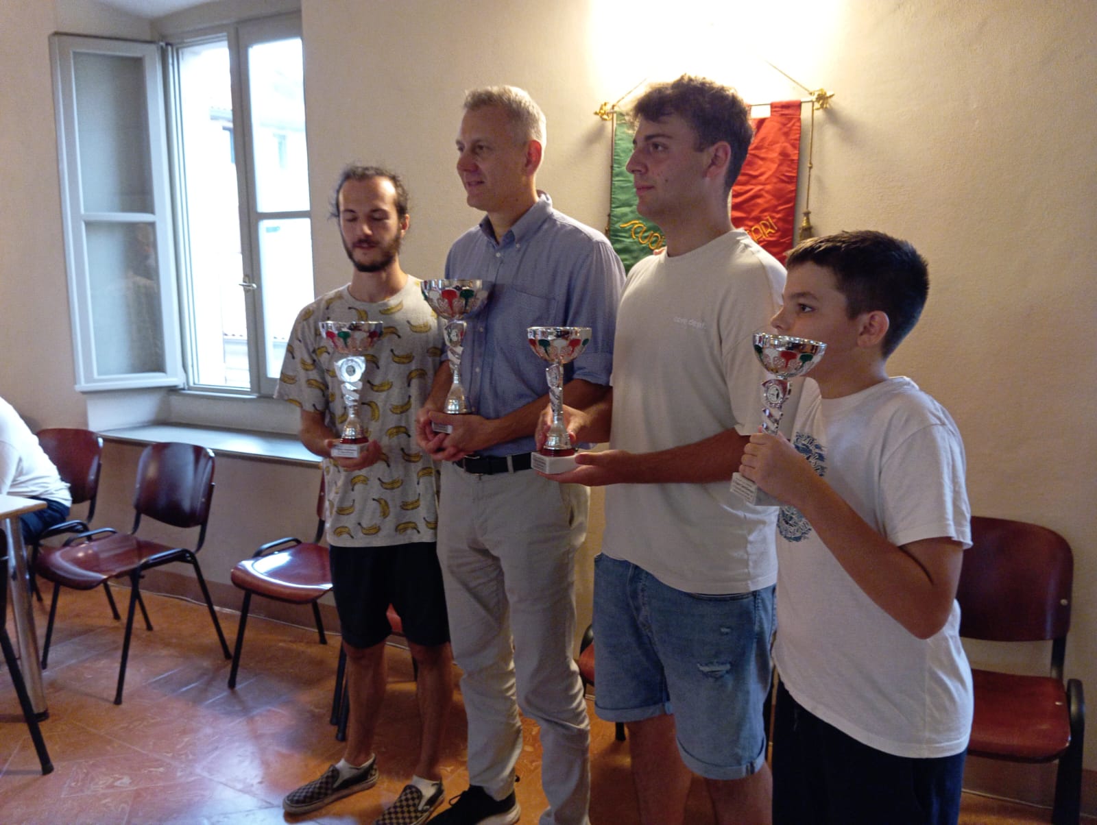Tre vincitori del torneo di scacchi mostrano i loro trofei insieme a un rappresentante del comune, all'interno di una sala consiliare luminosa.