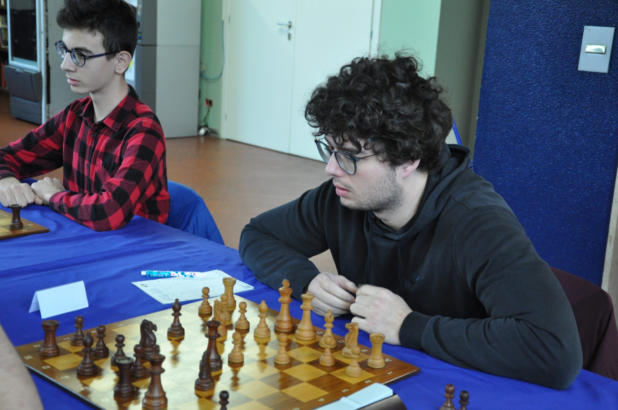 Two young men engaged in a game of chess at a table, with a chessboard displaying pieces in mid-game.