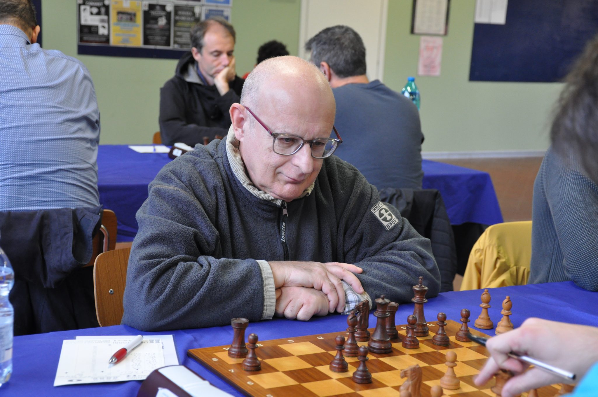 A focused individual with glasses seated at a chessboard during a chess game, with other players visible in the background.