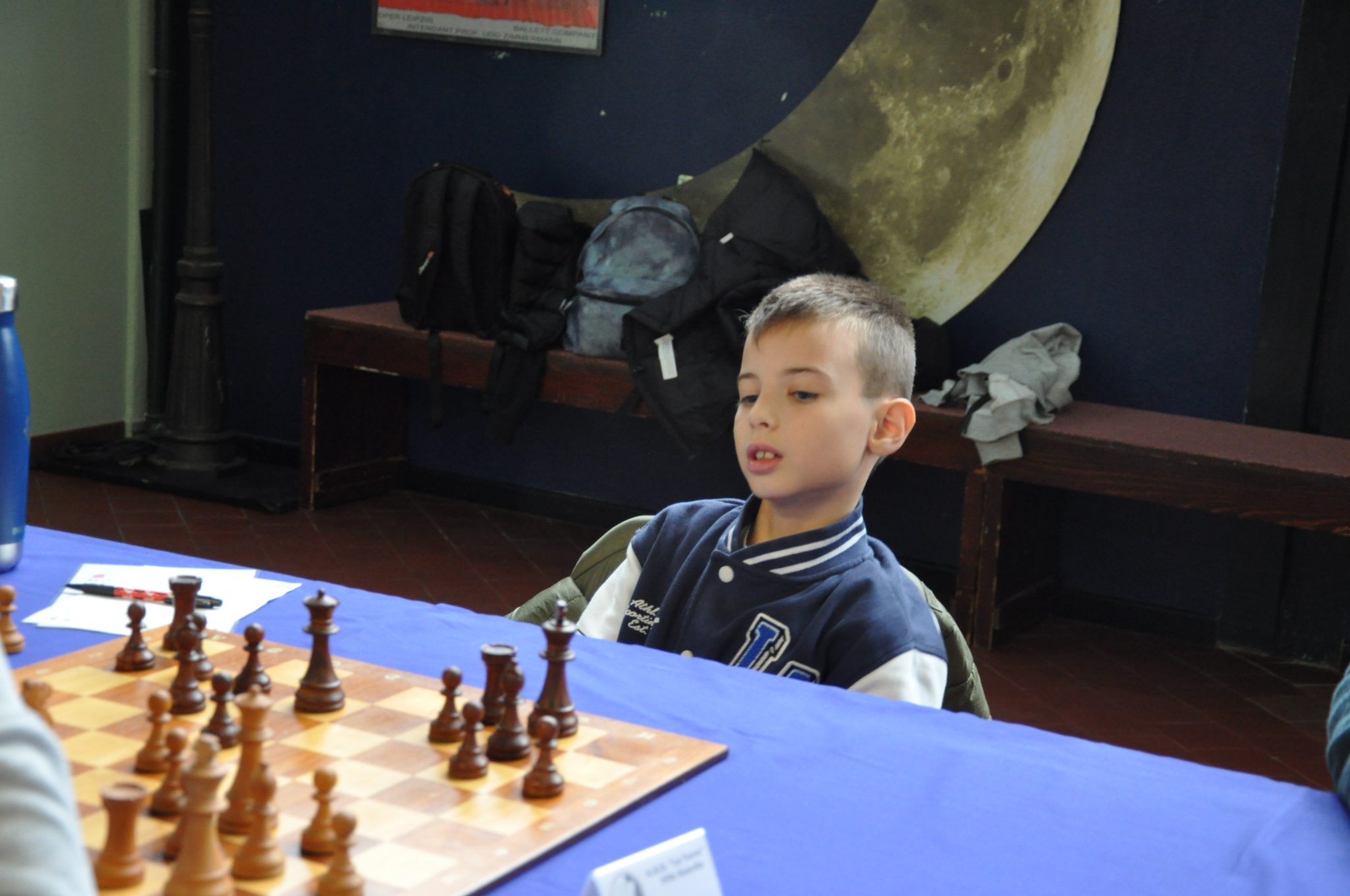A young boy with short hair, wearing a blue jacket, sits at a chessboard contemplating his next move, with a dark background and a large moon decoration behind him.