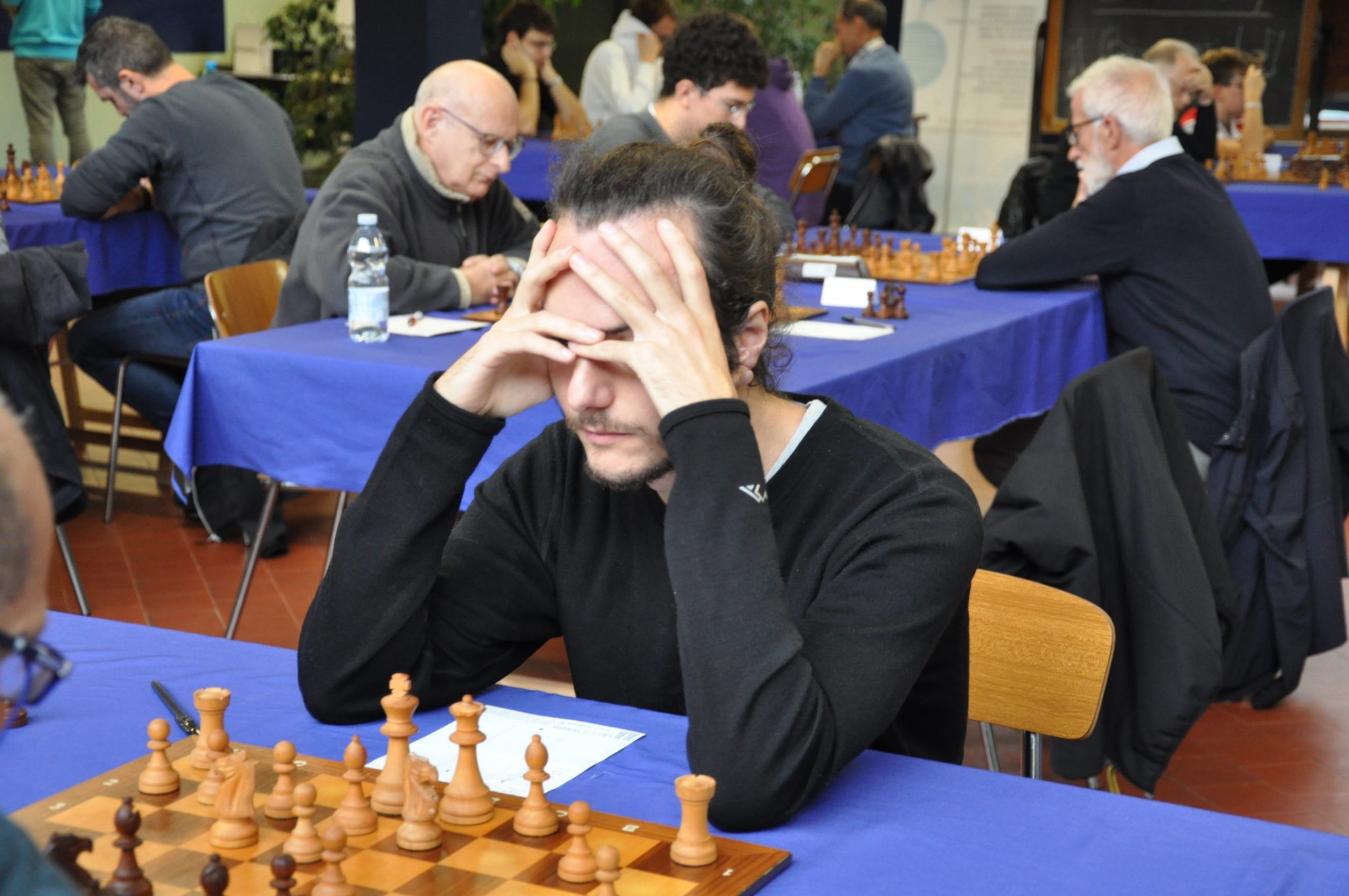 A chess player with long hair is deep in thought, resting his head in his hands while focusing on the game. In the background, other players are seated at tables with chessboards.
