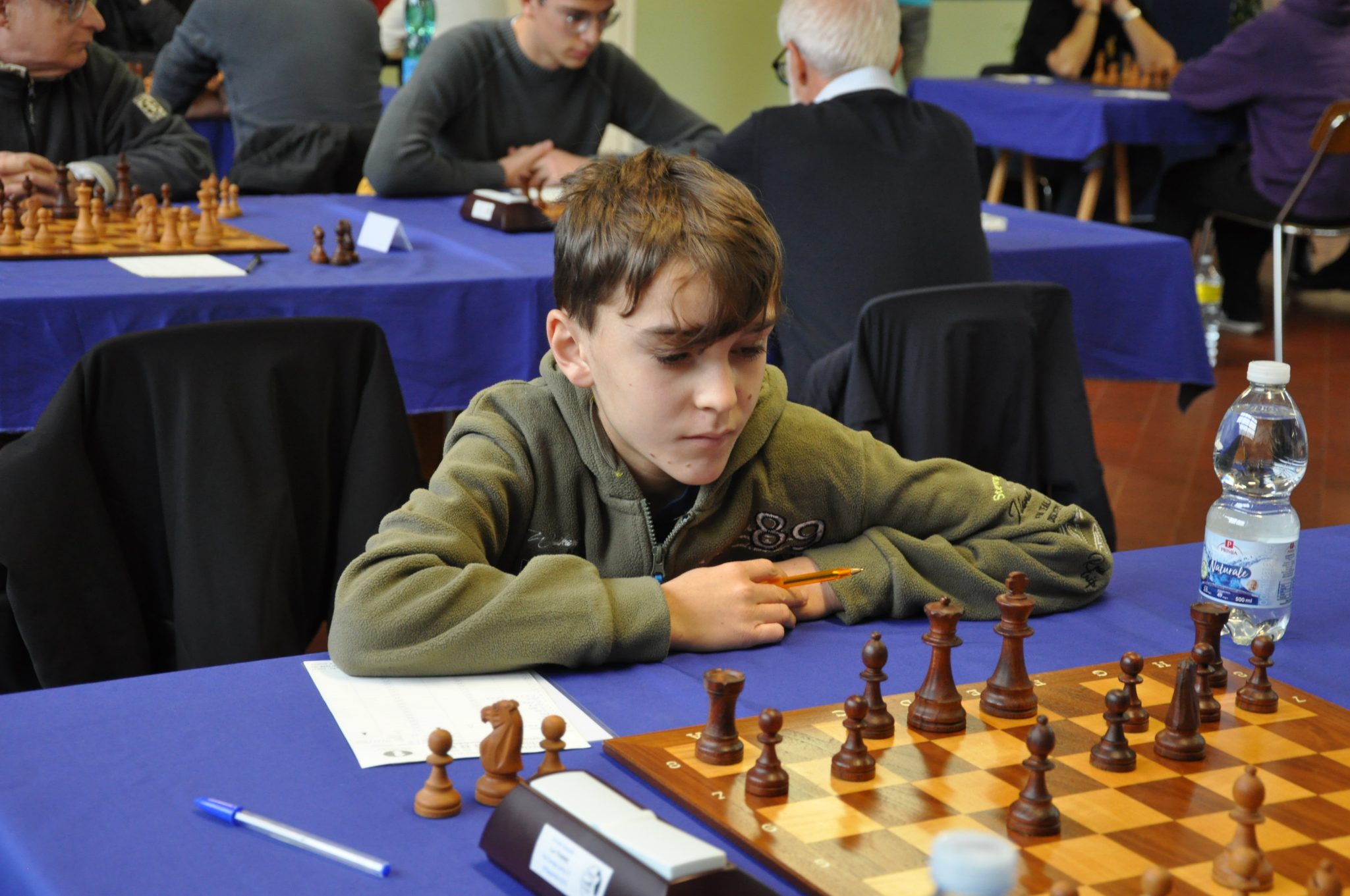 A young boy is deeply focused on a chess game, sitting at a table with a chessboard and pieces. Other players can be seen in the background.