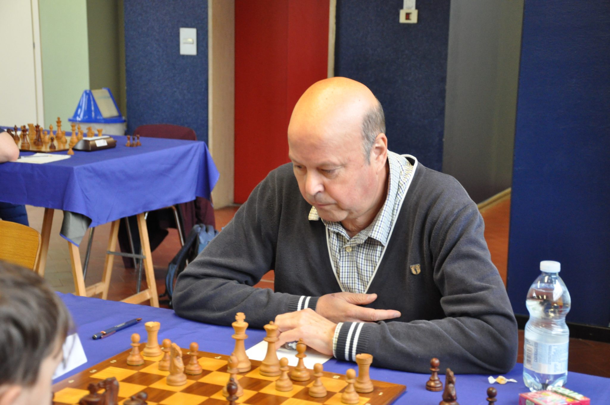 A focused individual is sitting at a chess table, contemplating a move during a game. The table is covered with a chessboard and pieces, and there are water bottles and other chess-related items in the background.