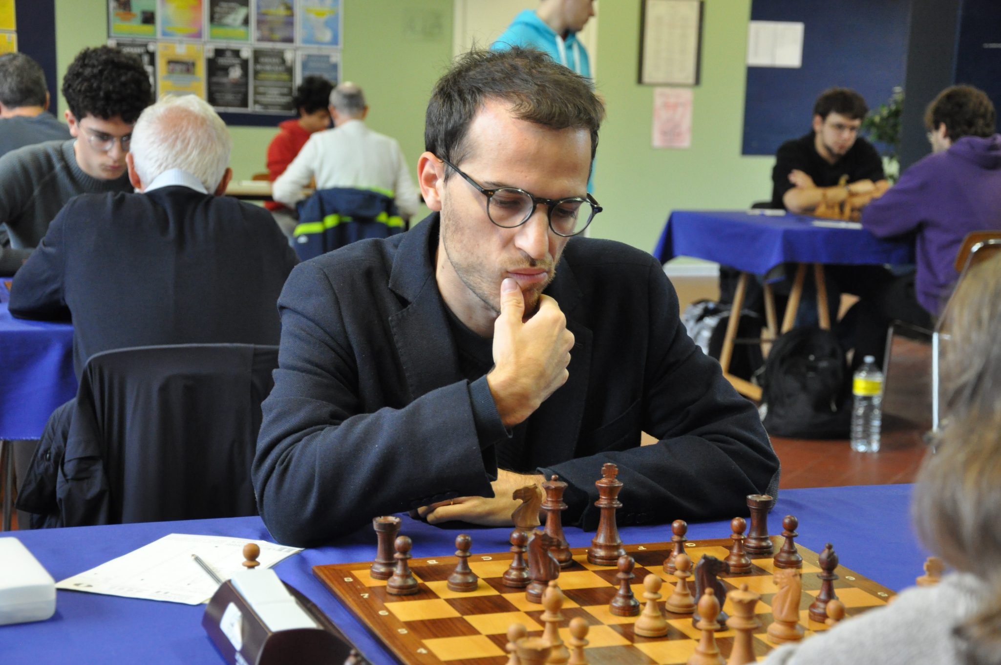 A chess player, deep in thought, is contemplating his next move during a tournament. The scene features multiple tables with other players engaged in the game.