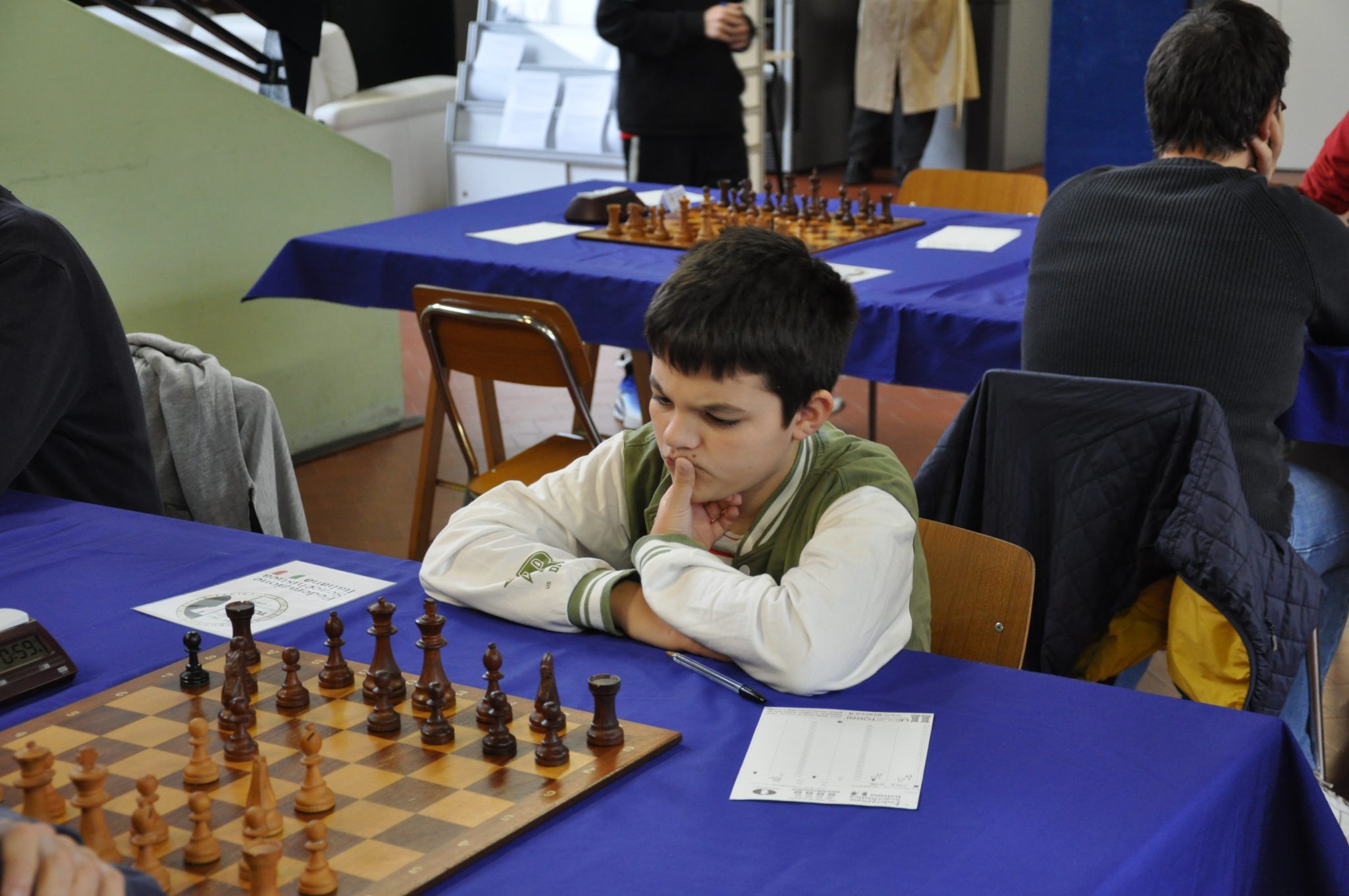 A young boy thoughtfully focused on a chess game at a competition, with a chessboard and pieces in front of him.