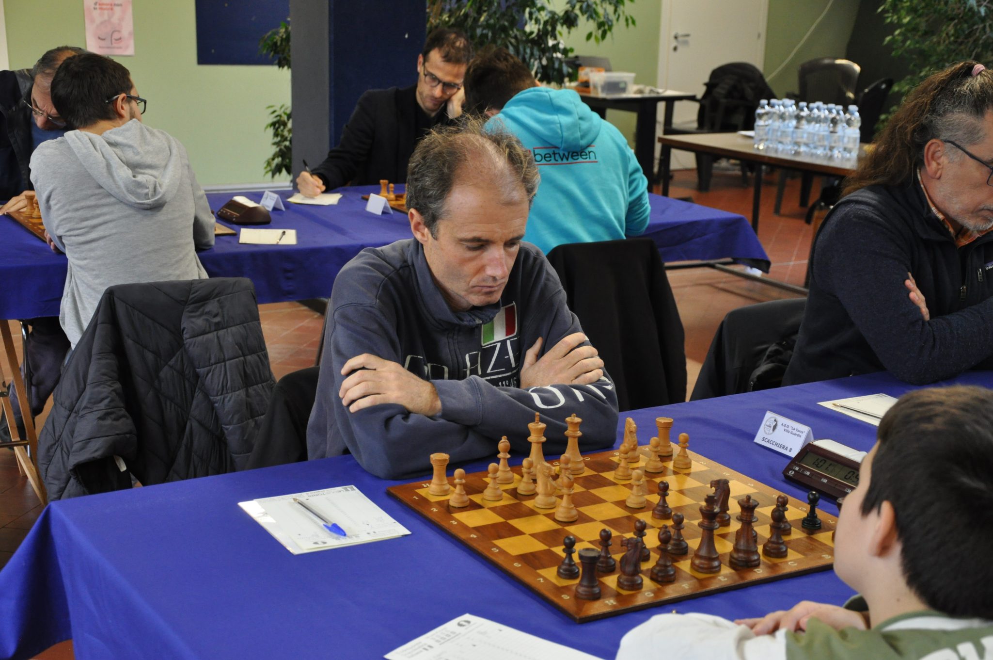 Chess tournament with a player concentrating on the board; other participants visible in the background.