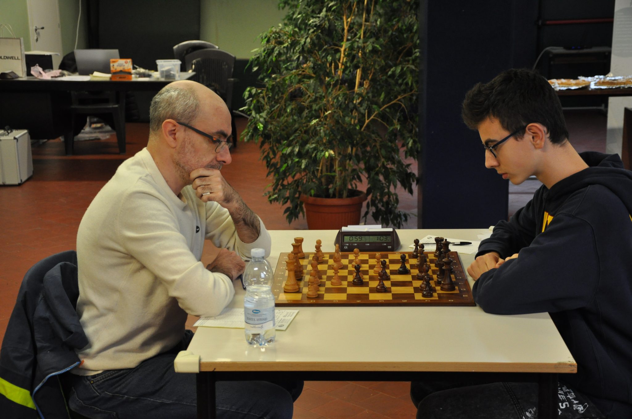 Two players engaged in a chess match, concentrating intently on the board, with a plant in the background.