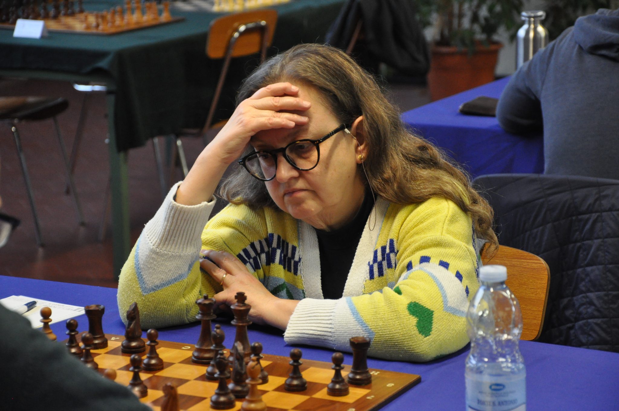 A focused female chess player in a yellow and green cardigan, contemplating her next move while resting her forehead on her hand. A chessboard is set up in front of her.