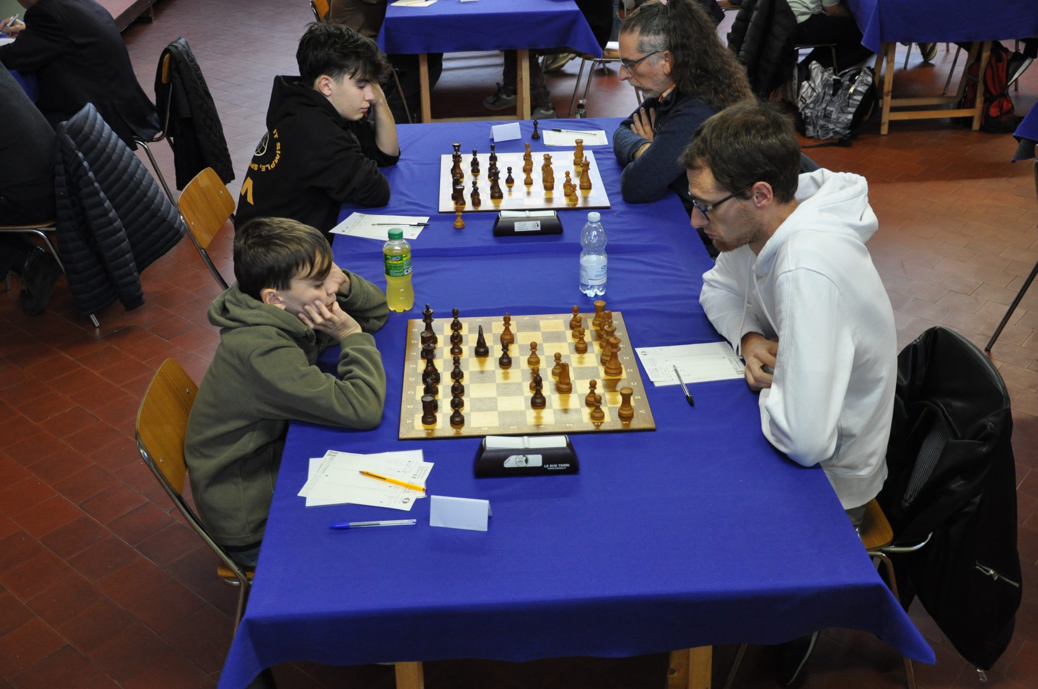 Two pairs of chess players engaged in a match, seated at tables with blue cloths. The players are focused on their games, with chessboards displaying pieces in various positions.