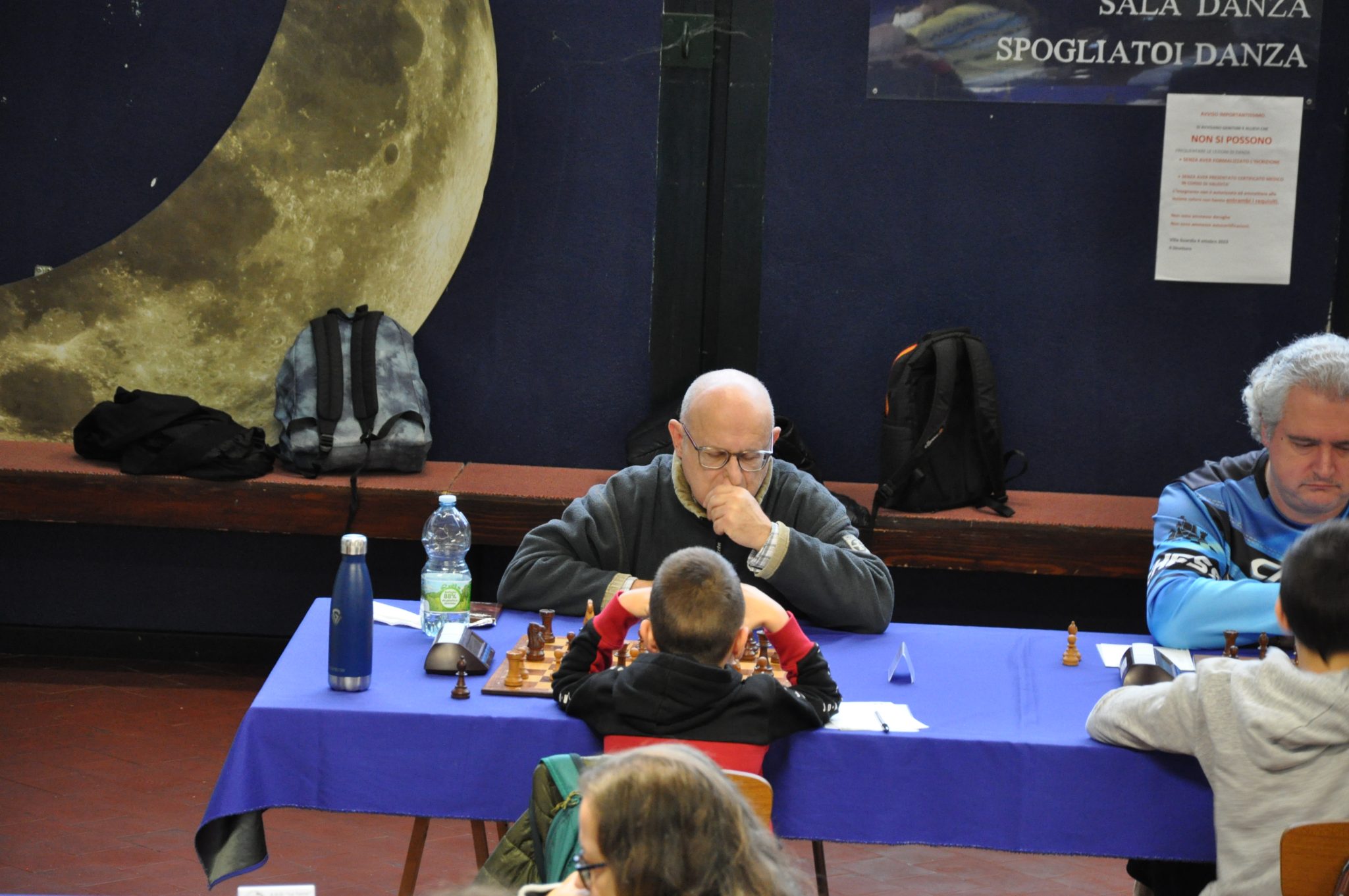 A chess competition featuring an elderly man deep in thought as he plays against a child, with a blue tablecloth and game pieces in front of them.