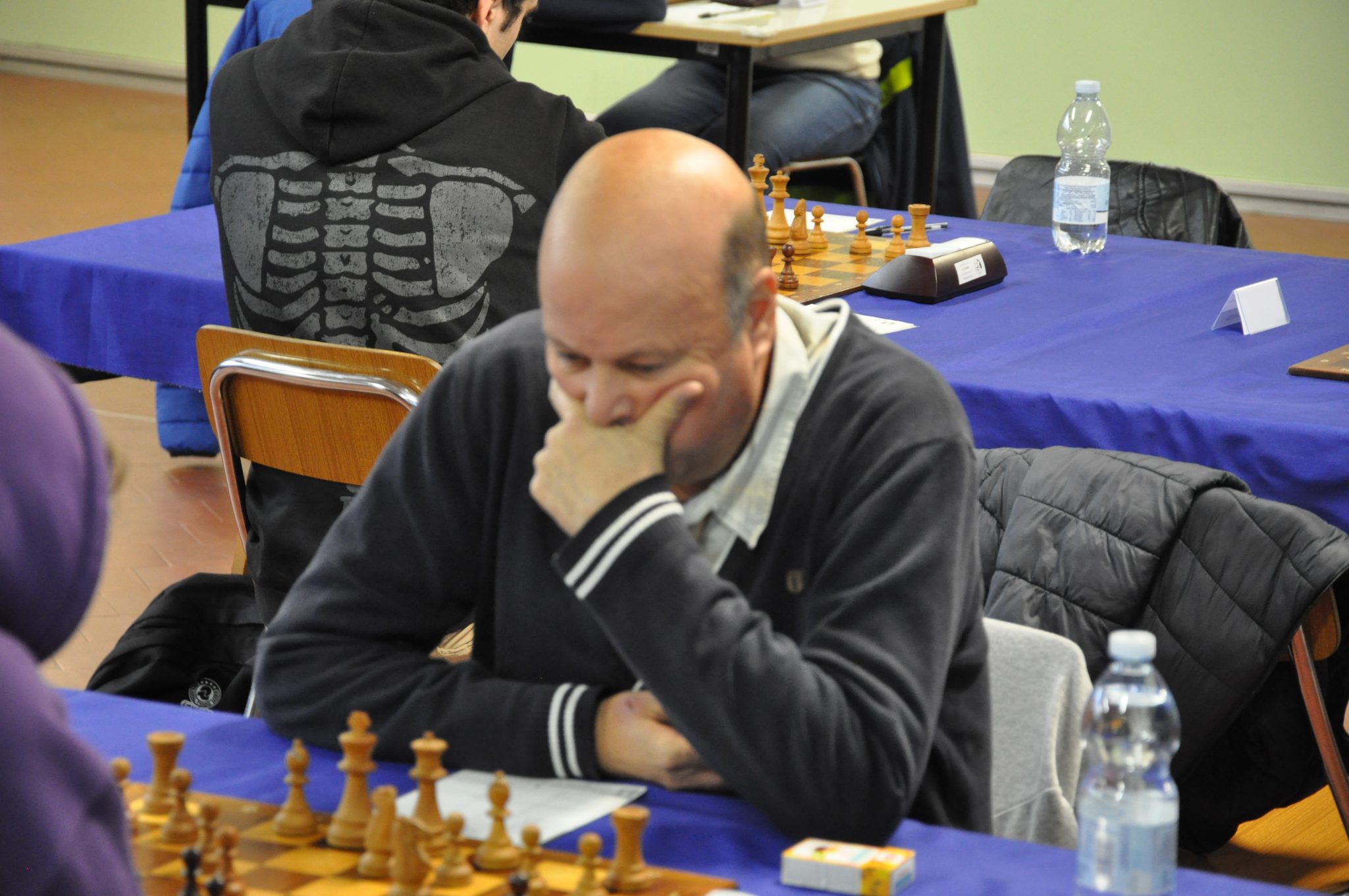 A focused individual with a shaved head is contemplating a chess move at a tournament, surrounded by chess pieces and opponents in the background.