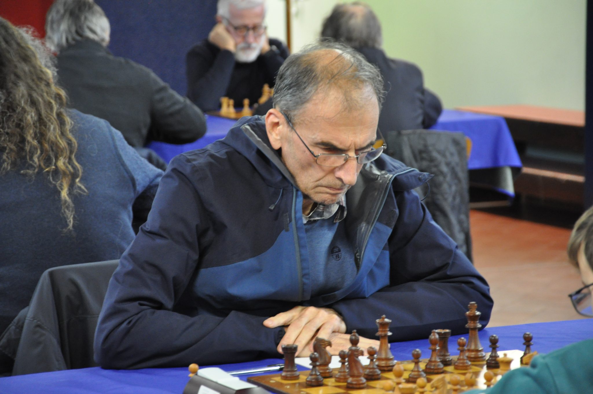 A focused chess player contemplating their next move during a match, with several chess pieces on the board and other players in the background.