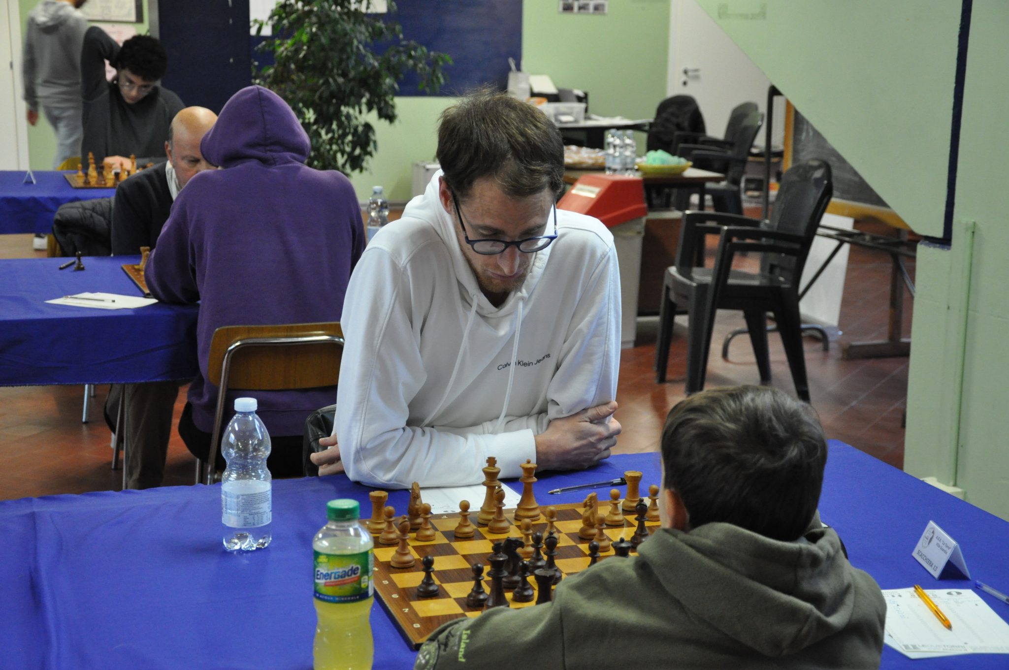Two chess players engaged in a match at a table, with a chessboard and pieces set up between them. One player is wearing a white shirt and appears focused, while the other is wearing a hooded sweatshirt.