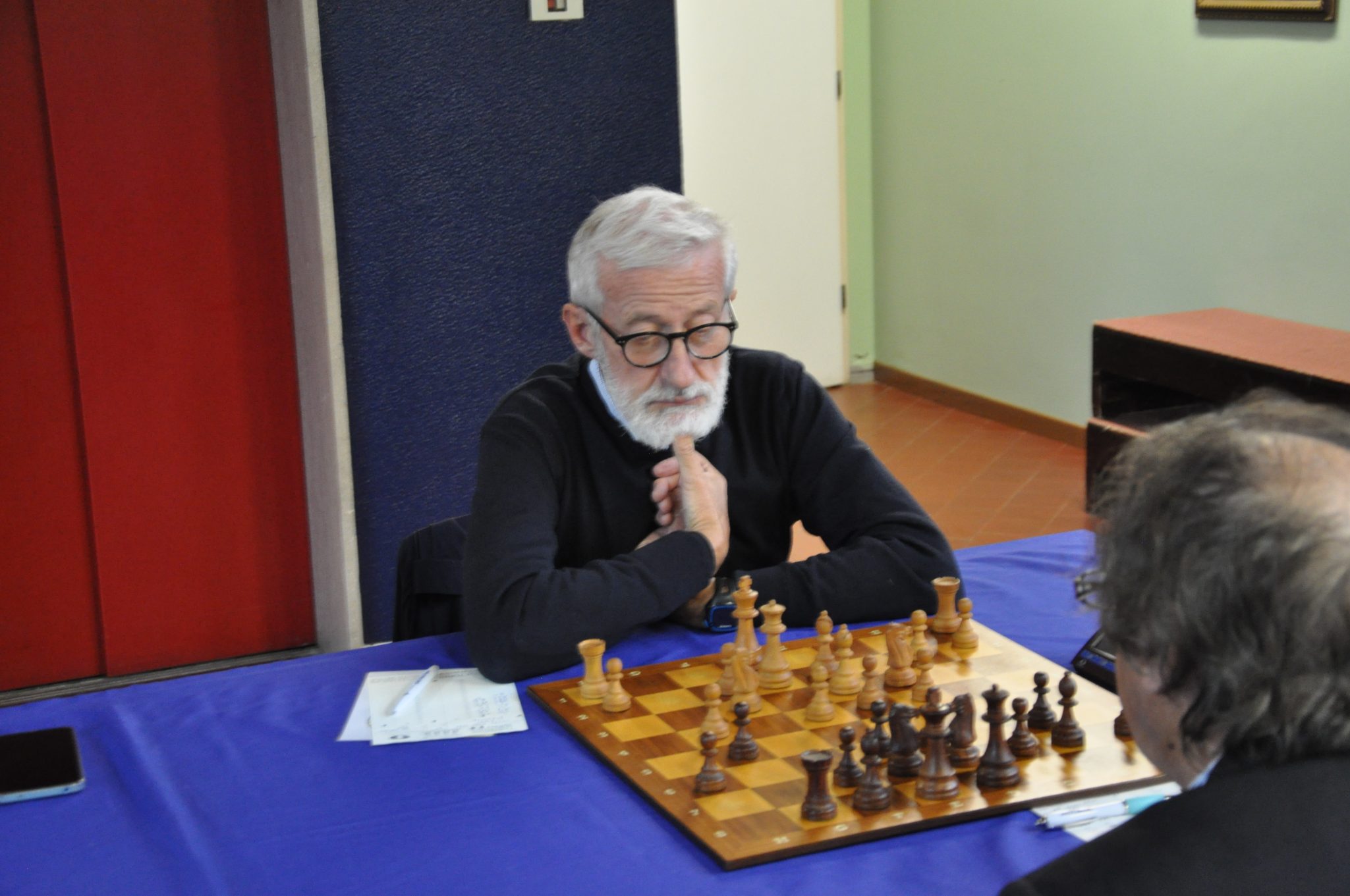 A serious-looking man with gray hair and a beard, sitting at a chessboard, contemplating his next move.