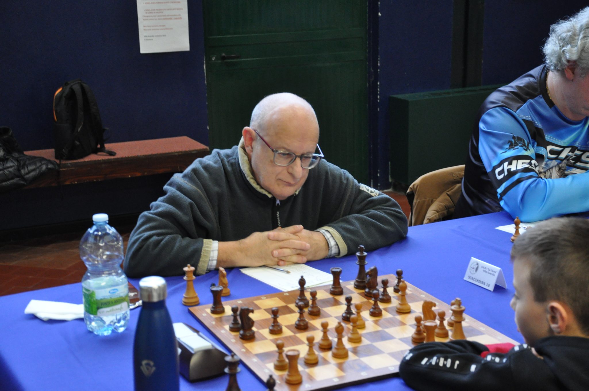 An older man concentrating on a chess game, sitting at a table with a blue tablecloth and a chessboard in front of him. A water bottle and other chess-related items are visible on the table.
