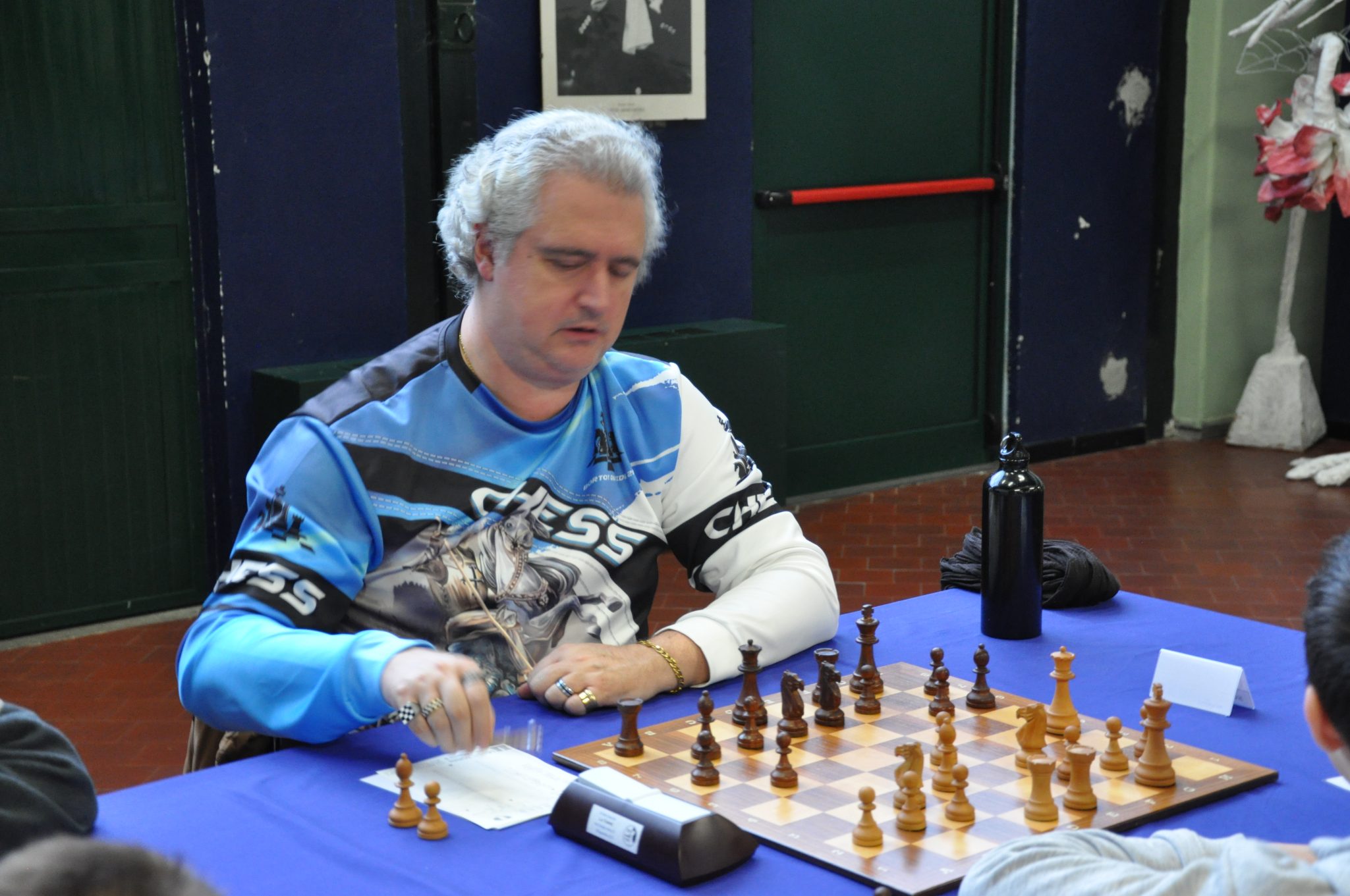 A person with gray hair playing chess at a table, wearing a blue chess-themed shirt, focused on the board.
