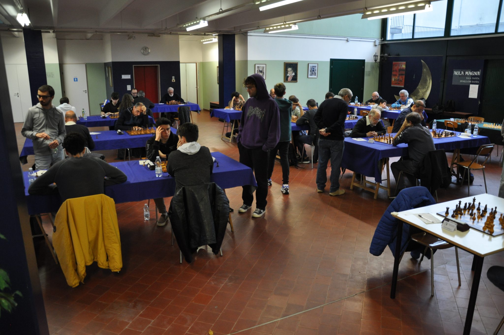 A spacious room filled with people engaged in chess games at tables covered with blue cloth.