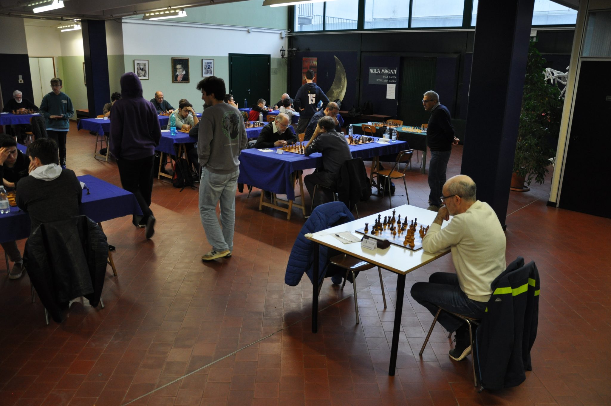 A chess tournament taking place indoors, with multiple participants seated at tables, some playing chess and others engaging in discussions or observing.