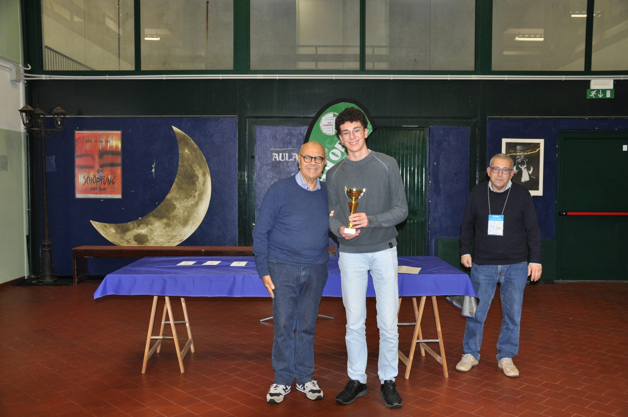 Two individuals, one holding a trophy, standing in front of a table with awards. The background features a moon decoration and a wall with posters.