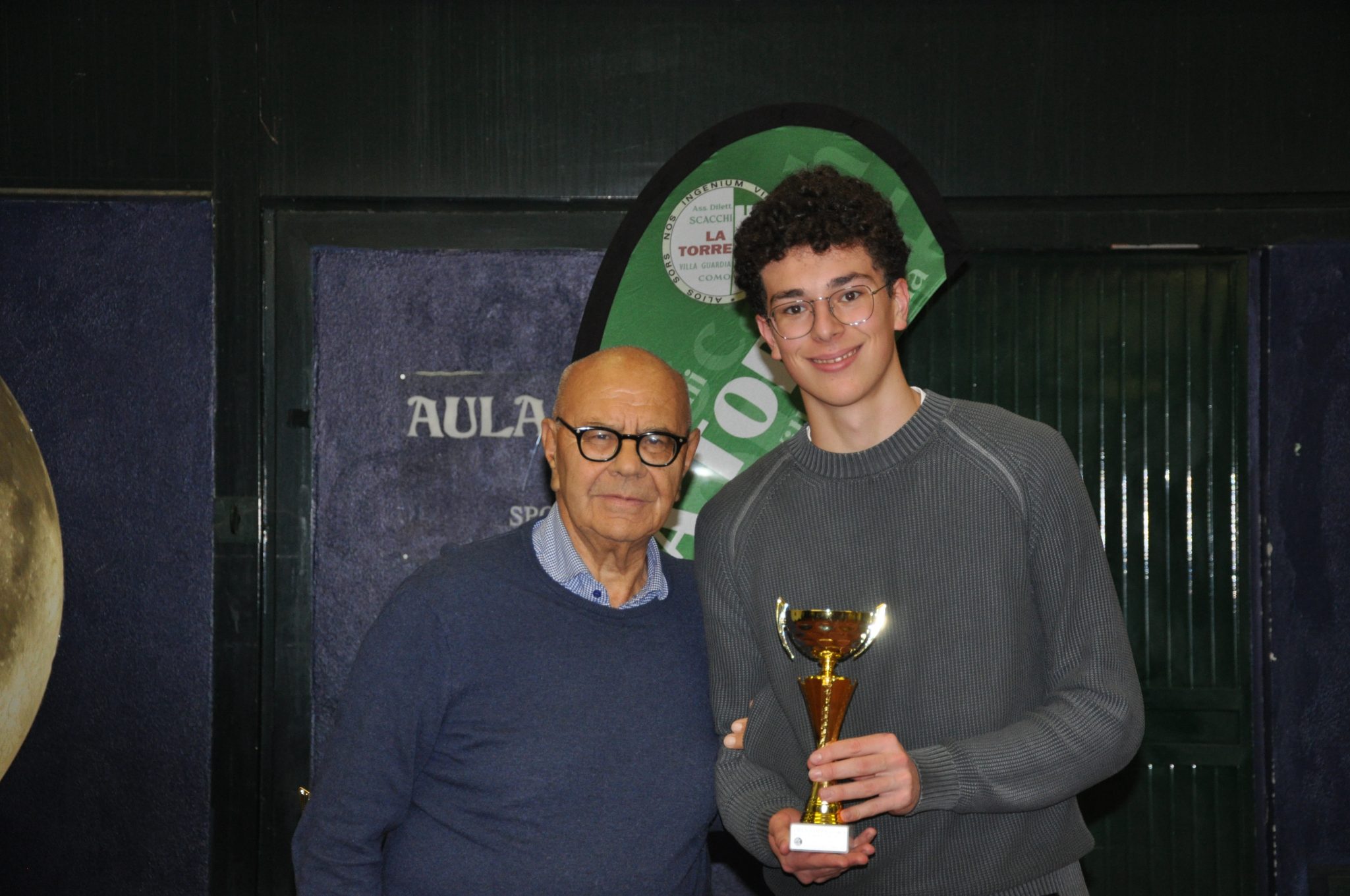 Two people standing together, one older man wearing glasses and a blue sweater, and a younger man holding a trophy, both smiling in front of a dark green backdrop.