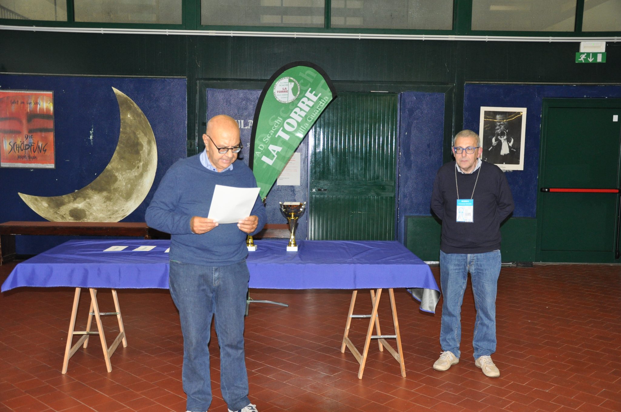 Two men standing in a room during an event, with one man reading from a paper and the other listening. A green banner and a trophy are visible on a table.