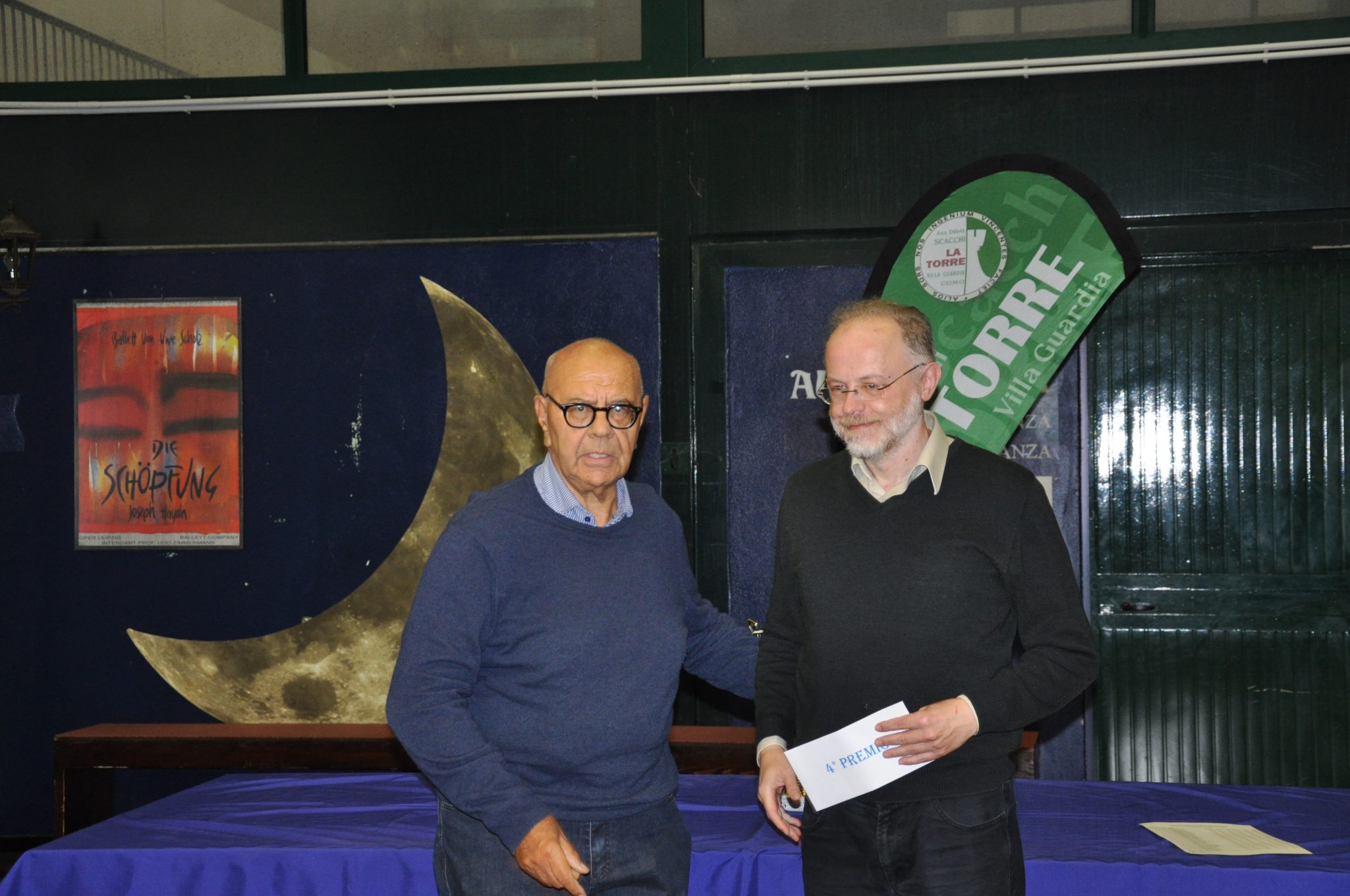 Two men in a formal setting, one handing a document or check to the other, with a moon-shaped decoration in the background.