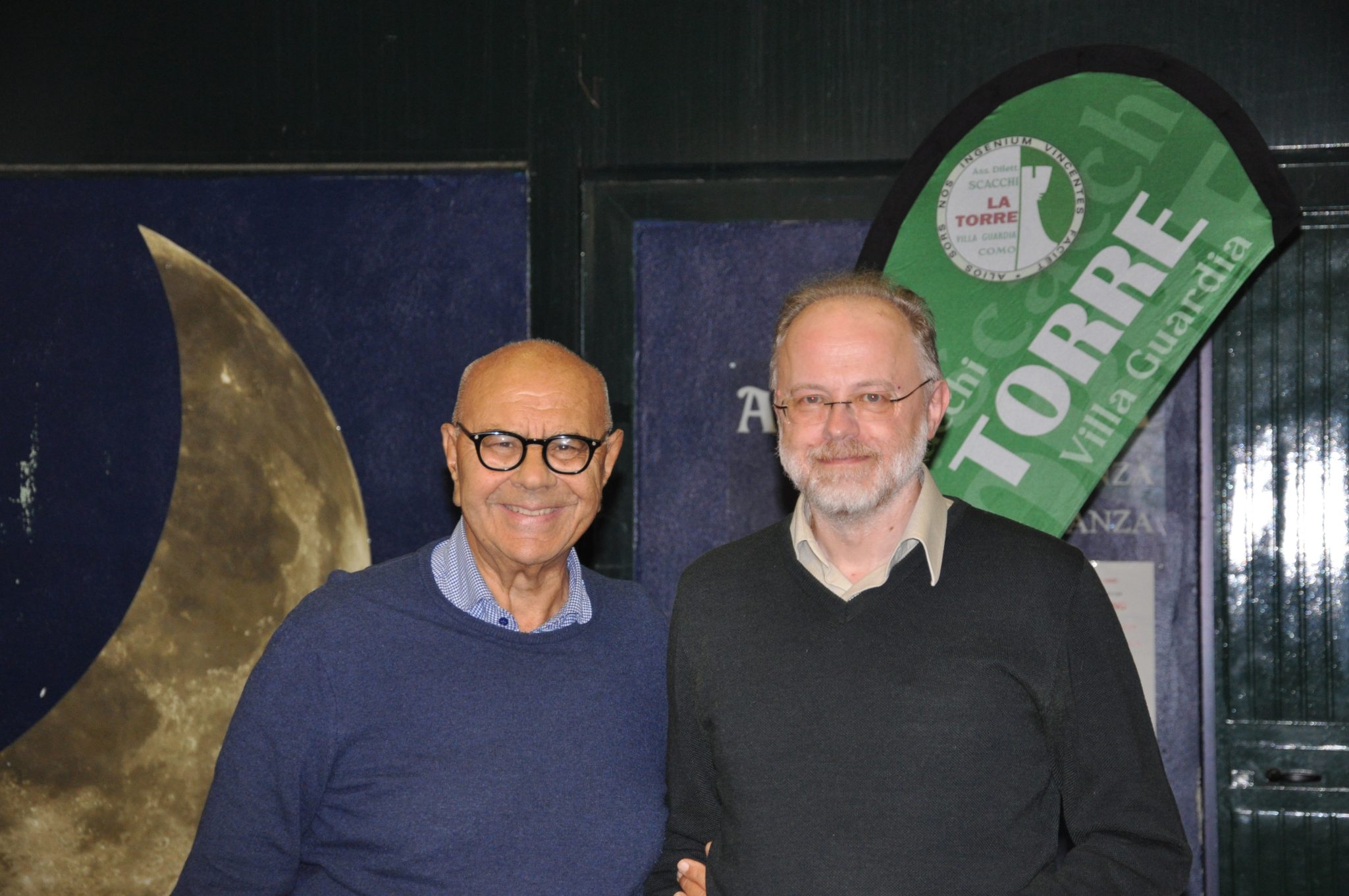 Two men smiling together in a venue, with a large crescent moon and a green sign in the background.