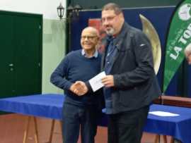 Two men standing side by side, one shaking hands with the other, in a formal setting with a table and decorative backdrop.