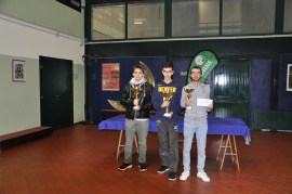 Three young men standing in an indoor setting holding trophies. The background appears to have a table with a blue cloth and a banner.