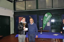 A young man in a black leather jacket holds a trophy while posing for a photo with an older man in spectacles, who is wearing a blue sweater. Background includes a green banner and tables set up for an event.