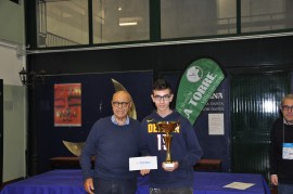 Two individuals standing at an indoor event, one holding a trophy while the other presents a certificate, with banners in the background.